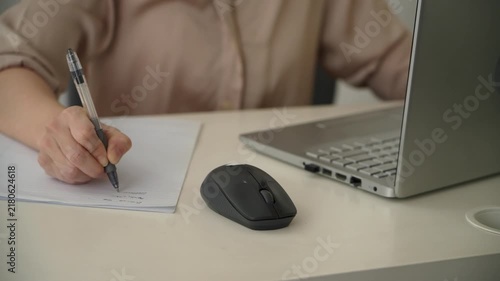 Close up of a woman writing with a pen on paper beside a laptop and wireless mouse at an office desk. Everyday note taking, planning and office work.