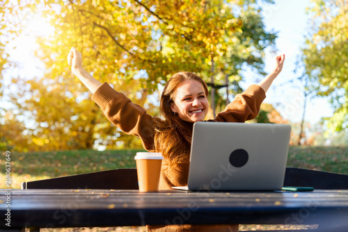 Joyful adult student enjoying sunny autumn day with laptop and coffee outdoors.