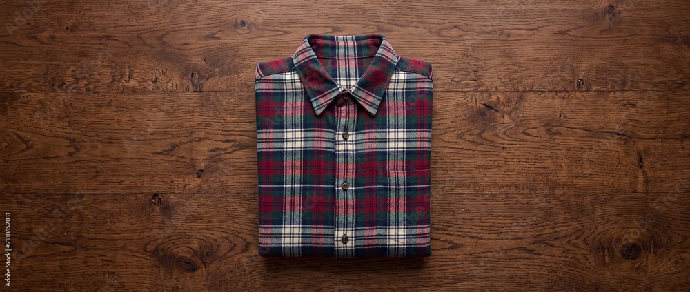 Fototapeta premium Folded plaid flannel shirt with classic red green and blue checkered pattern resting on dark rustic wooden surface for cozy autumn apparel and fashion themes.