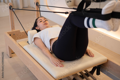 Asian woman performing Pilates on a reformer with feet in straps, building core strength and flexibility through controlled movement for fitness, balance, mobility, wellness, and body awareness.