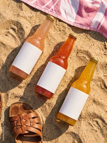 Three colorful beverage bottles with blank labels resting on golden sand next to a sandal and striped towel, evoking a summer beach day.