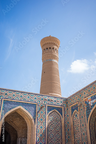 Fotografie Historic Islamic architecture in Bukhara, Uzbekistan, featuring a tall decorative brick minaret rising above blue and turquoise tiled arches