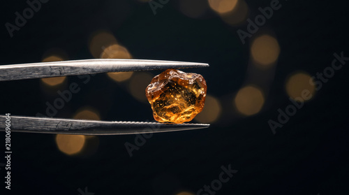 Tweezers holding a raw orange gemstone against a dark background