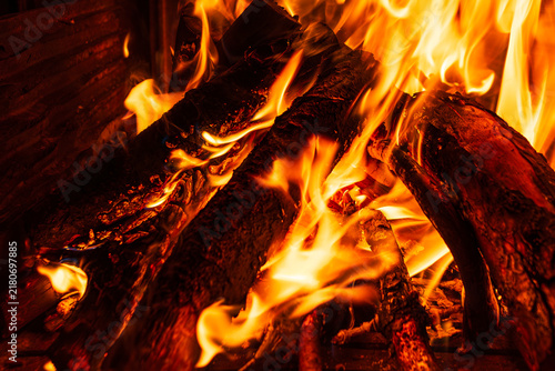 Close up of burning firewood logs with bright orange flames and glowing red embers inside a rustic stone fireplace, warm cozy winter background of a wood fire at night