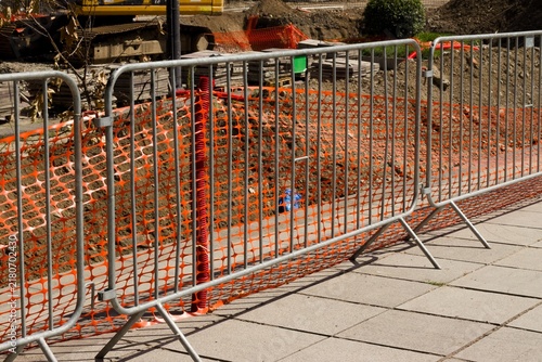 Canvastavla Urban roadwork area with temporary metal barriers, orange safety mesh, construction materials and excavation equipment beside a pedestrian sidewalk
