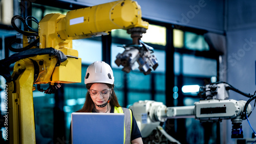 female robotics engineer uses a laptop to program an artificial intelligence system. She is in an R and D lab  testing and controlling multiple industrial robotic arms.