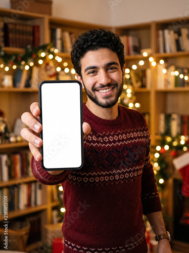 happy young man shows smartphone with white blank screen mockup on background of festive christmas garland lights and decorative shelves in cozy evening home room interior