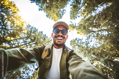 Happy young man taking selfie in a forest - Cheerful male traveler walking through a scenic green grove on a sunny day