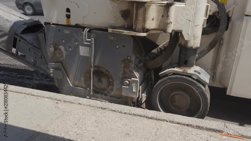 Close up view of an asphalt milling machine drum cutting and removing old road surface next to a concrete curb during pavement renovation.