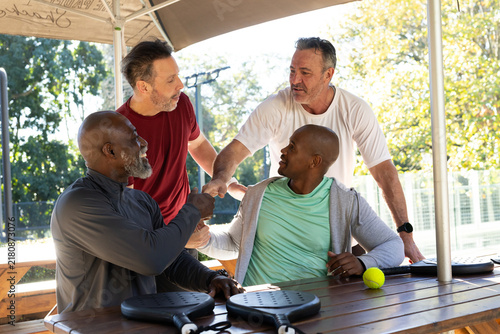 Diverse men in gym gear shaking hands, chatting at picnic table under shade with paddles, ball