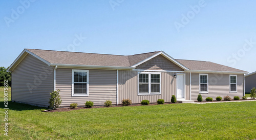 Exterior view of single story manufactured modular house with beige vinyl siding and front steps set on spacious green grass lawn under clear sky