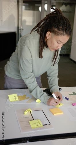Vertical video: Nonbinary African American leaning in writing notes on mockups for design at office