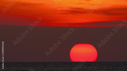 Giant bright red sun setting over calm ocean horizon with vibrant crimson clouds sky. Extreme telephoto static shot of dramatic sunset dusk over water.