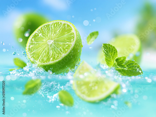A sliced fresh green lime rests splashing in clear blue water, paired with vibrant mint leaves, whole limes blurred in soft‑focus tropical background.