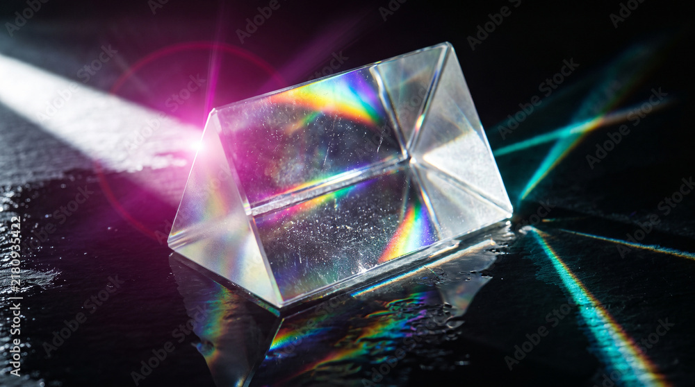 Obraz premium Triangular crystal prism lying on wet dark glossy surface refracting white light beam into rainbow color spectrum. Physics optics experiment, bright lens flare reflection.