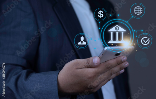 Obraz na plátně Online mobile banking and global money transfer concept with businessman hand holding smartphone displaying glowing bank icon and connected network nodes on dark background