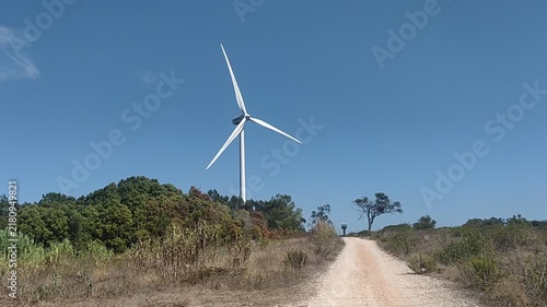A majestic wind turbine rises beside a winding dirt road under a bright blue sky, evoking a deep sense of hope for a sustainable future. Nobody is in this peaceful landscape.