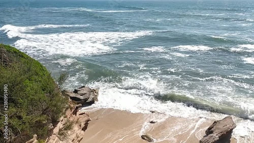 A serene highangle view of powerful ocean waves crashing onto a secluded sandy beach with rugged, vegetated cliffs. This wild landscape evokes raw natural beauty. Nobody.