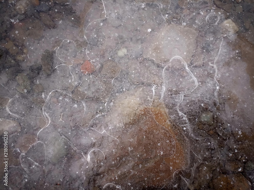 Top down close up view of a frozen puddle showing detailed ice crack patterns, air bubbles, and ground beneath.
