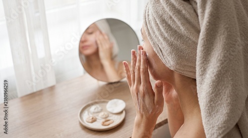 Woman applying facial skincare cream during morning beauty routine with towel on head in front of mirror, healthy skin moisturizing and self care treatment at home vanity background