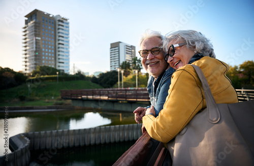 Smiling senior couple leaning on a railing in a city park, enjoying the view of a pond and cityscape, embracing their golden years together