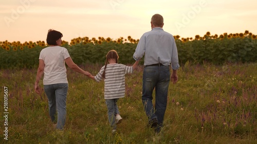 Family mother father man woman parent walking through field with child sunflowers at sunset, Happy family time girl child outdoors, Parents holding hands with child, Sunset walk in countryside in