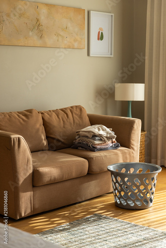 Brown two-seat sofa is holding folded linen near window, grey laundry basket casting cutout shadows