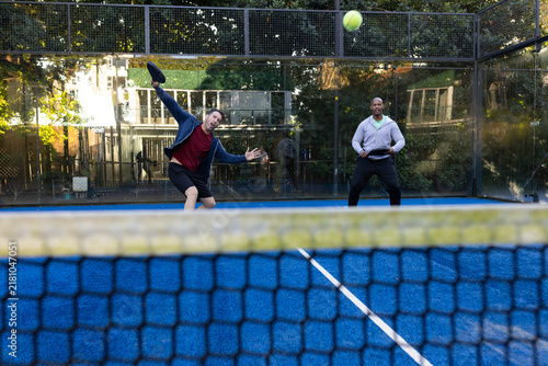 Diverse male friends playing paddle on blue court, swinging paddles, watching yellow ball near net