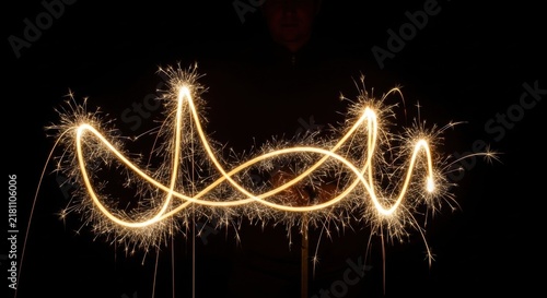 Long exposure photography of golden sparkler light trails forming a wavy abstract pattern against a solid black background, captured at night with bright sparks and glowing curves.