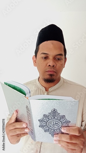 Bild auf Leinwand A Muslim man wearing a traditional black cap and beige attire is reading the Qur