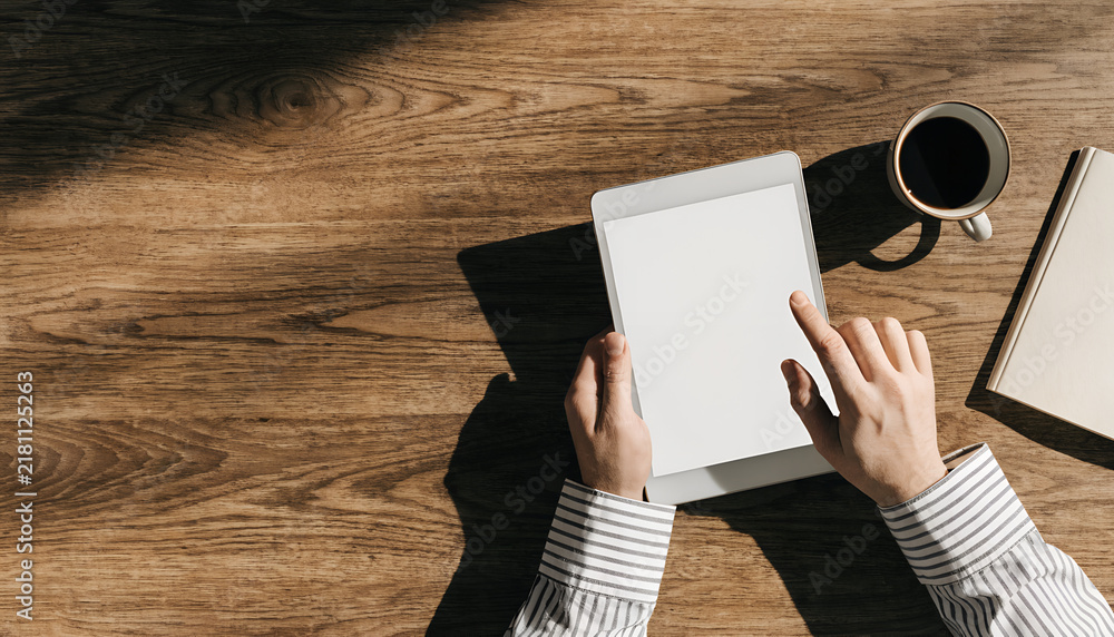 Obraz premium A person using a blank tablet on a wooden table with a cup of coffee and a notebook nearby, illuminated by sunlight.