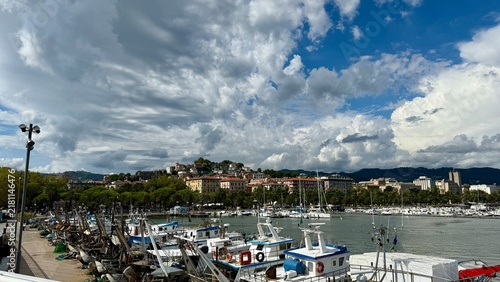 Fishing boats and small vessels moored in the marina of La Spezia, Italy, along the Ligurian coast. Dramatic clouds fill the sky above the waterfront, with  the town in the background. 