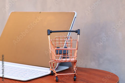 Miniature shopping cart in front of an open tablet with blank mockup screen on wooden table, online shopping and digital commerce concept.