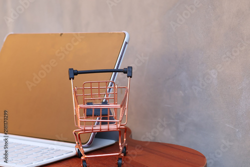 Miniature shopping cart in front of an open tablet with blank mockup screen on wooden table, online shopping and digital commerce concept.