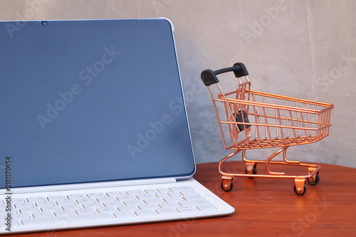 Miniature shopping cart next to a modern tablet with blank mockup screen on a wooden table, online shopping and e-commerce business concept with copy space.