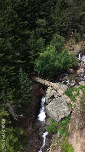 Dos montañistas cruzando un puente sobre un río de alta montaña vistos desde un dron 4K en toma cenital, rodeados de naturaleza salvaje y paisaje alpino