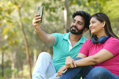 Indian happy young couple sitting on green grass in a beautiful park and taking a selfie together with a smartphone.