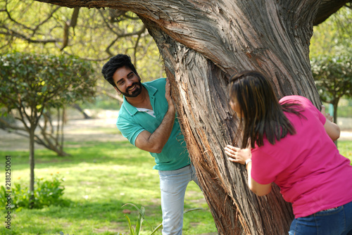 Quadro su tela Indian happy young couple playfully looking at each other from behind a tree in a beautiful green park, sharing a joyful romantic moment surrounded by nature