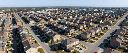 Aerial view of a newly built suburban housing development with uniform townhouses and paved streets under clear blue sky. Illustration for real estate investment articles or urban