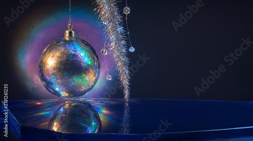A close-up of a holographic Christmas bauble with iridescent surface reflecting rainbow light on a dark blue reflective surface. Illustration for holiday design, festive packaging,