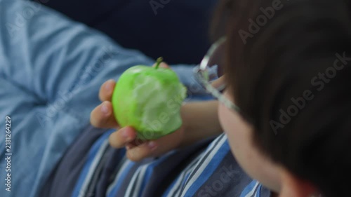 cute young boy with glasses eating a fresh green apple. Healthy childhood nutrition, daily vitamin snack, and casual indoor lifestyle portrait of a kid enjoying organic fruit.
