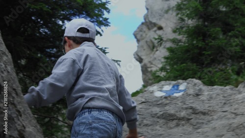 Young boy hiker in a white cap climbing rocks and enjoying a beautiful panoramic mountain view. Active childhood, summer trekking adventure, and outdoor nature exploration concept.