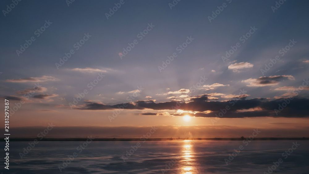 Fototapeta premium Sunset over frozen lake surface with clouds and sun rays beaming.