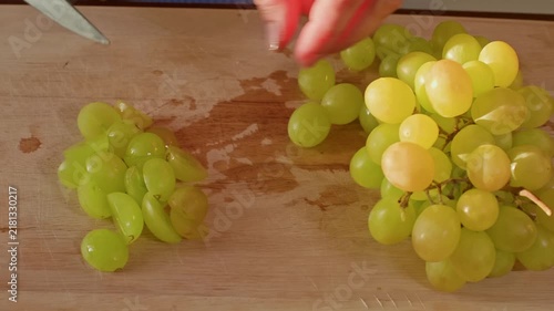 Person uses knife hand to divide fresh white grape bunches on wood surface, close up static shot capturing food preparation, sorting action in natural kitchen lighting.