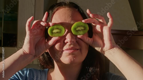 Woman holds two kiwi fruit slices over eyes like eyeglasses, playful pose captured with natural window lighting, minimal camera movement in bright interior setting.
