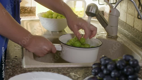 Person rinsing fresh green grapes in white colander at kitchen sink, bright natural light streaming through window, fruit preparation close up.