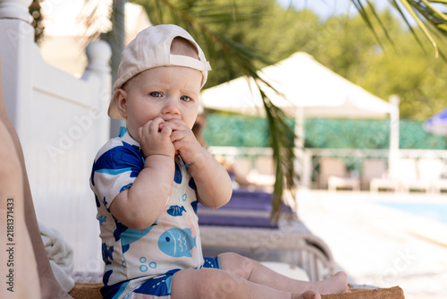 Cute baby in swimwear sits by the pool with hands near mouth