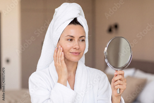 Beautiful woman in a bathrobe and towel on her head looking at herself in a hand mirror