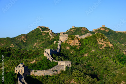 Foto The Jinshanling Great Wall, an ancient stone fortification, winds across lush green mountain slopes under a vivid blue sky, with steep stone steps and aged watchtowers rising amid dense vegetation
