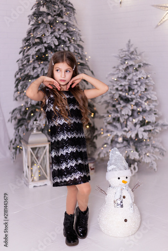 Elegant Little Caucasian Girl in Sequin Dress Posing Near Snowy Christmas Trees with Snowman and Star Decor